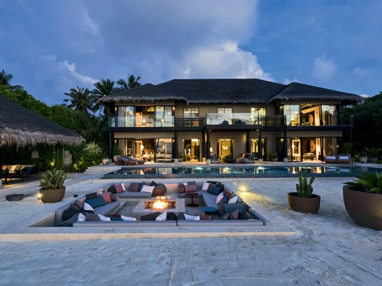 Two-storey Velaa Private Residence lit at dusk with a long pool and a sunken fire-pit lounge on the sand, Noonu Atoll, Maldives