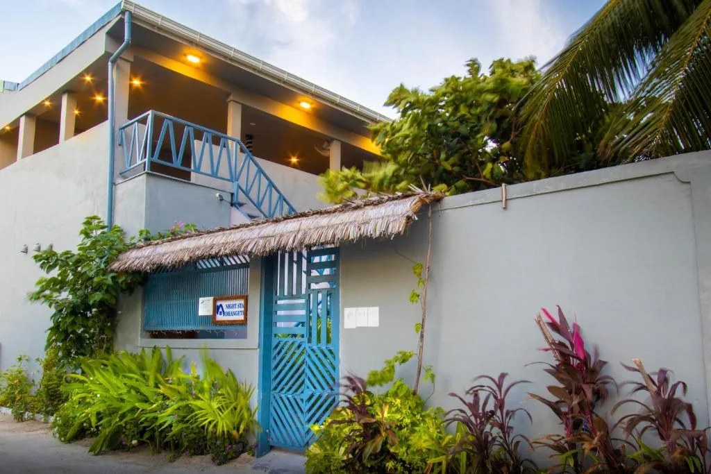Two-story guesthouse with blue-railed balcony, thatched-roof gate, tropical garden and sign reading 'Night Sunset' at Night Star Dhangethi, Dhangethi, Maldives