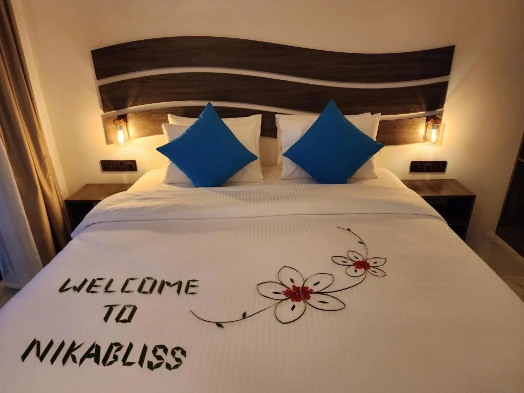King bed with blue pillows, towel runner reading 'WELCOME TO NIKABLISS', wooden headboard and wall sconces at Nikabliss, Dhigurah, Maldives