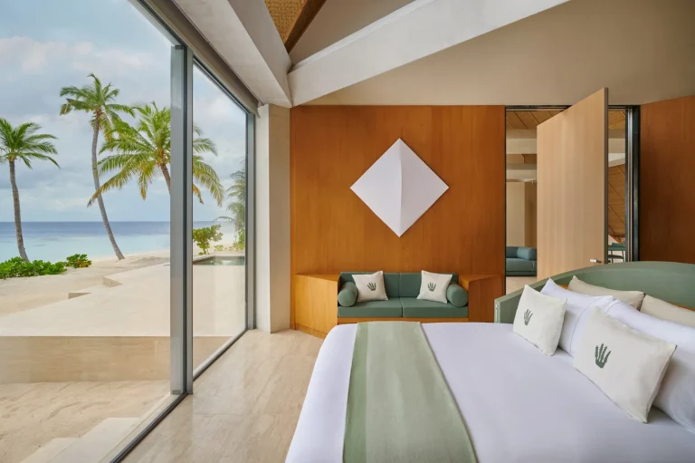 Bedroom with a white bed, green throw and a glass wall onto the beach and palms, Nilaa Coral Island, Maldives