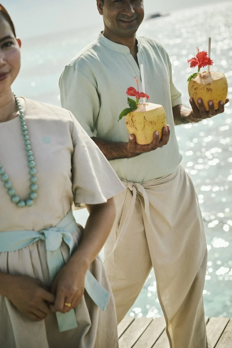 Two hosts in linen holding fresh coconut drinks by the sea, Nilaa Coral Island, Maldives