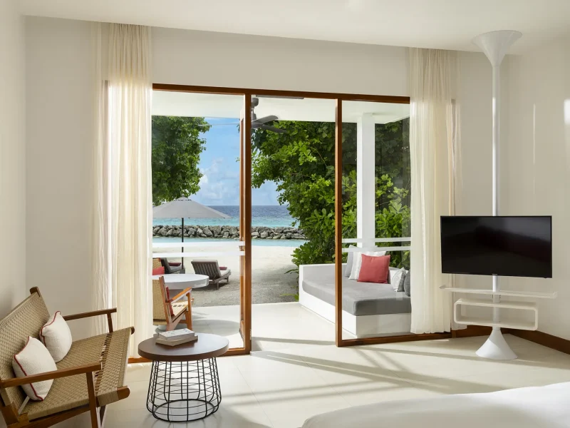 Bright bungalow room with a chair, side table and glass doors open to a terrace and the beach, Niva Dhigali Maldives