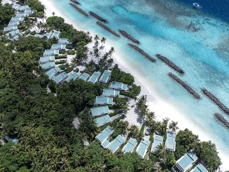 Overhead view of a row of white roofed bungalows among palms beside a white beach and turquoise water, Niva Dhigali Maldives