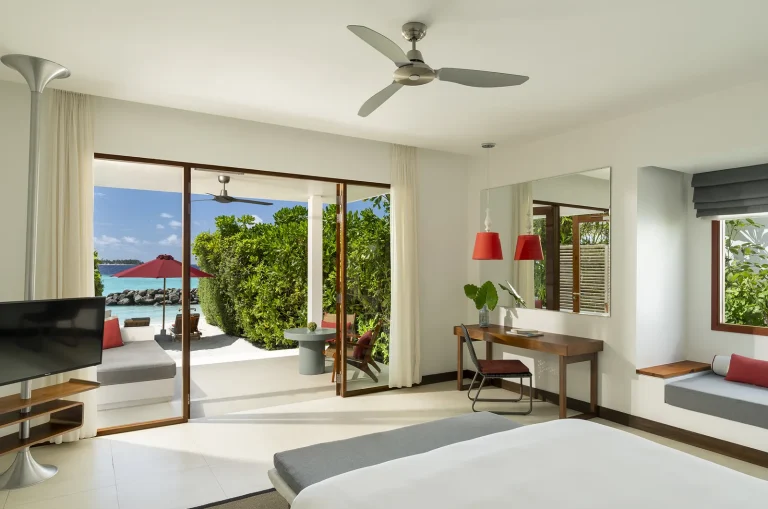 Bright bedroom with a king bed, desk, ceiling fan and doors open to a green garden and beach in a Beach Villa, Niva Dhigali Maldives