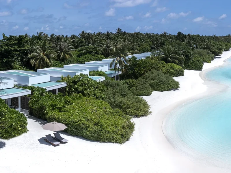 Aerial view of white beach villas tucked into dense green vegetation along a long white beach and lagoon, Niva Dhigali Maldives