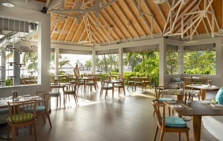 Bright open restaurant with timber tables, rattan chairs and a high timber ceiling at Capers, Niva Dhigali Maldives