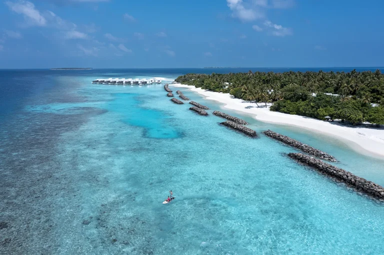Aerial view of a long green island edged by white sand with overwater villas stretching into a turquoise lagoon, Niva Dhigali Maldives