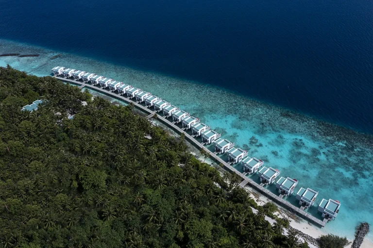 Aerial view of a long line of overwater villas along a jetty beside a green island and reef, Niva Dhigali Maldives