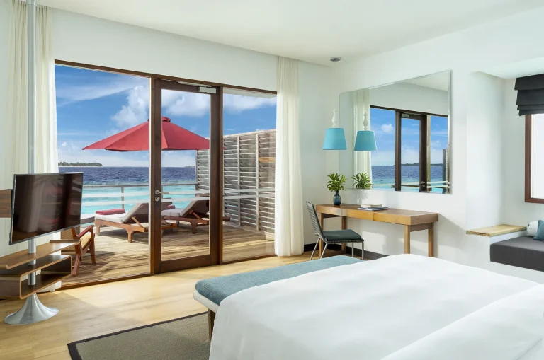 King bed facing glass doors onto a deck with a red umbrella and the lagoon in a Water Villa, Niva Dhigali Maldives
