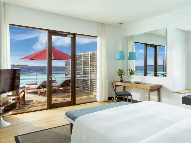 King bed facing glass doors onto a deck with a red umbrella and the lagoon in a Water Villa, Niva Dhigali Maldives