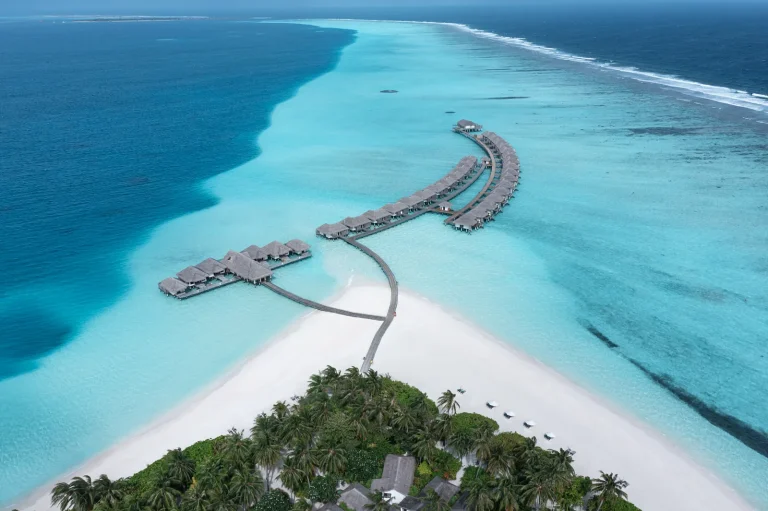 Aerial view of a long curved jetty leading over a pale turquoise lagoon to overwater villas and the restaurant at Niva Velassaru Maldives
