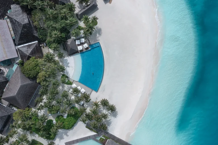 Overhead view of a curved blue infinity pool beside a wide white beach and lagoon with thatched buildings among trees, Niva Velassaru Maldives