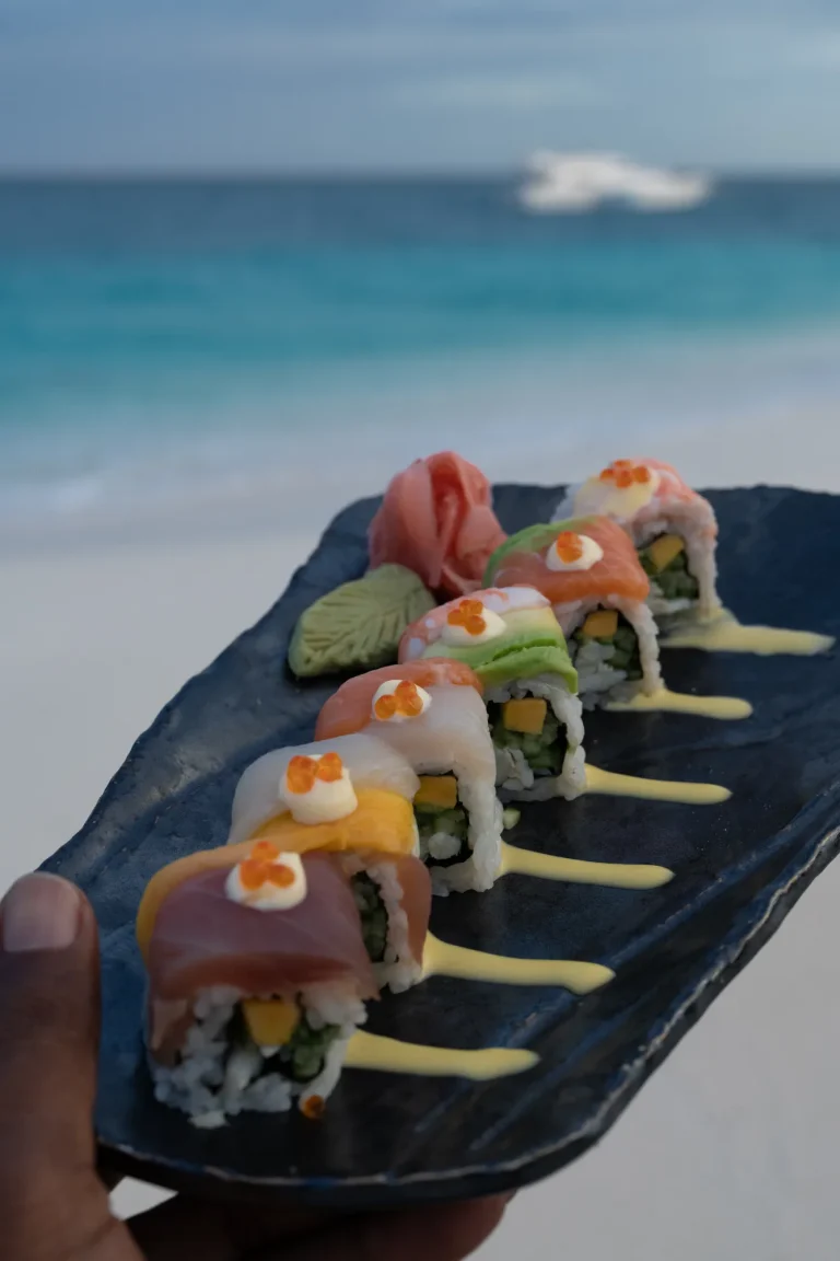 Plate of colourful sushi rolls held up against the beach and lagoon at Sora, Niva Velassaru Maldives