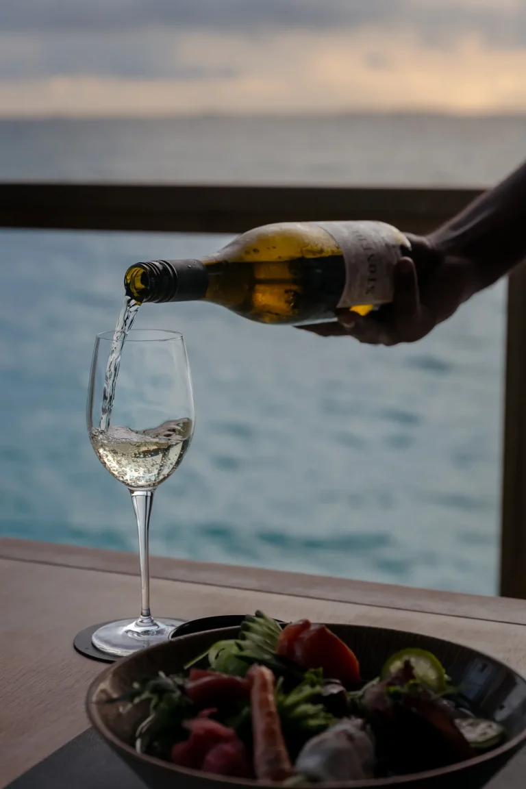 White wine being poured into a glass on a table overlooking the lagoon at sunset, Niva Velassaru Maldives