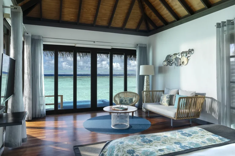 Bedroom and lounge with floor-to-ceiling glass doors onto a sun deck and turquoise lagoon in a Water Villa with Pool, Niva Velassaru Maldives
