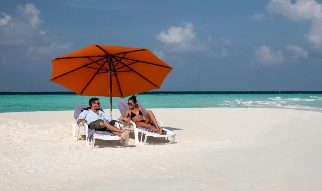Two people relaxing on white-sand beach under an orange umbrella, turquoise ocean and distant palm trees in the background at Old Town Inn, Gaafaru, Maldives