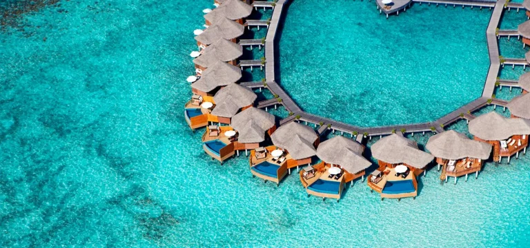 Aerial view of thatched Baros Water Pool Villas with private pools on a curved jetty over a turquoise lagoon, North MalΓ© Atoll, Maldives