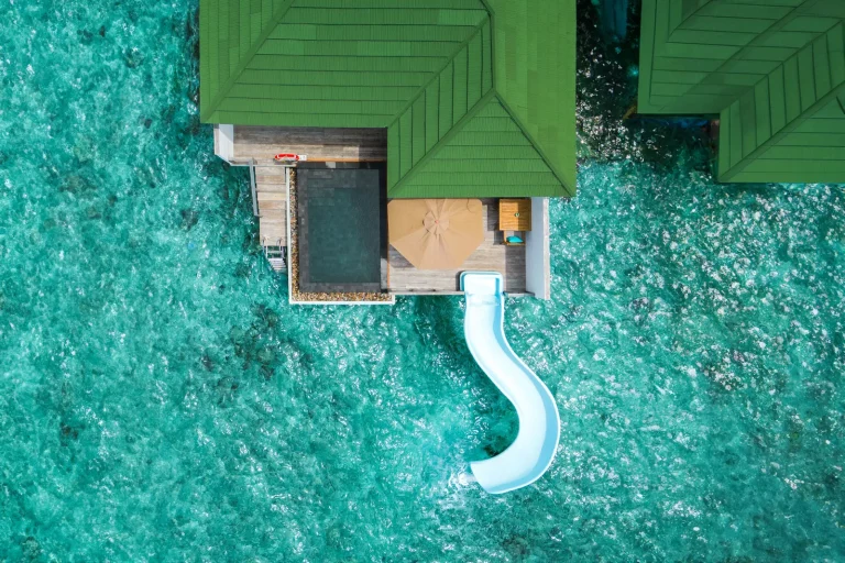 Aerial view of a green-roofed overwater villa with a private pool and a white slide curling into the lagoon at Siyam World Maldives, Noonu Atoll