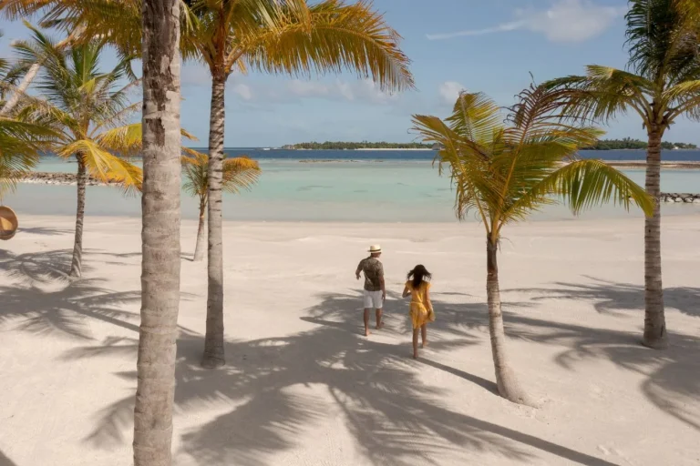 Couple walking under palm trees on a white sand beach with a calm lagoon at Patina Maldives, Fari Islands