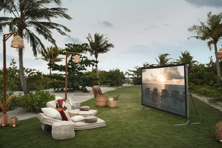 Outdoor cinema screen on a lawn with cushioned loungers under palm trees at Patina Maldives, Fari Islands