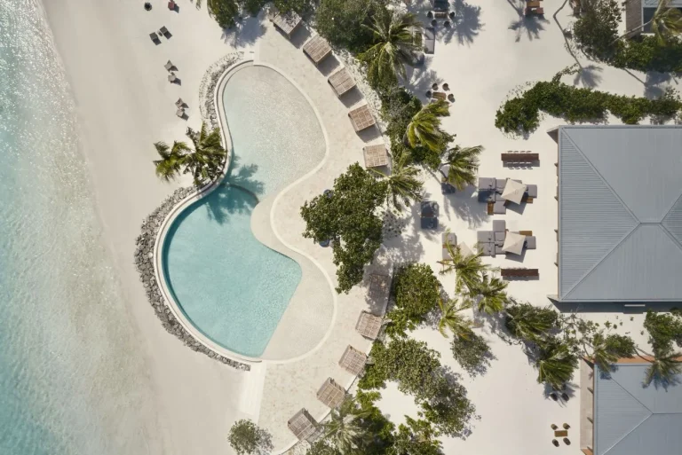 Overhead view of a curved swimming pool with sun loungers beside the beach at Patina Maldives, Fari Islands