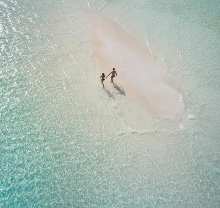 Aerial view of two people walking on a pale sandbank in shallow turquoise water near Patina Maldives, Fari Islands