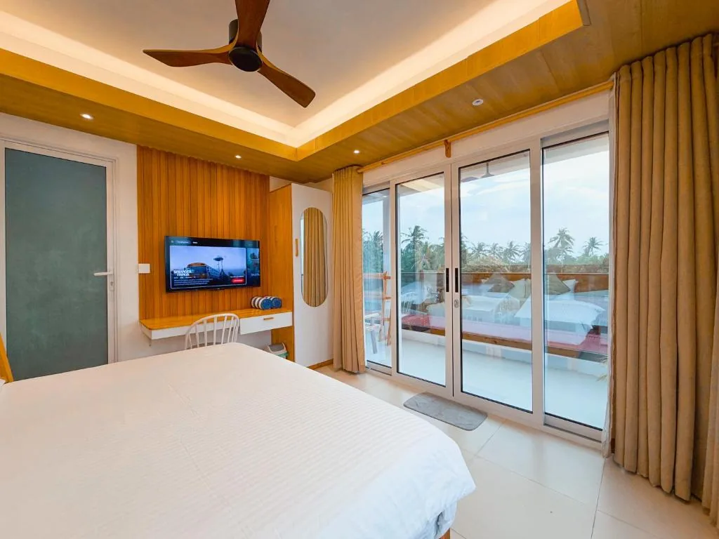 King bed with wood-paneled headboard, wall TV, desk and sliding glass doors to balcony with palm-tree view at Pavona Maldives, Gaafaru, Maldives