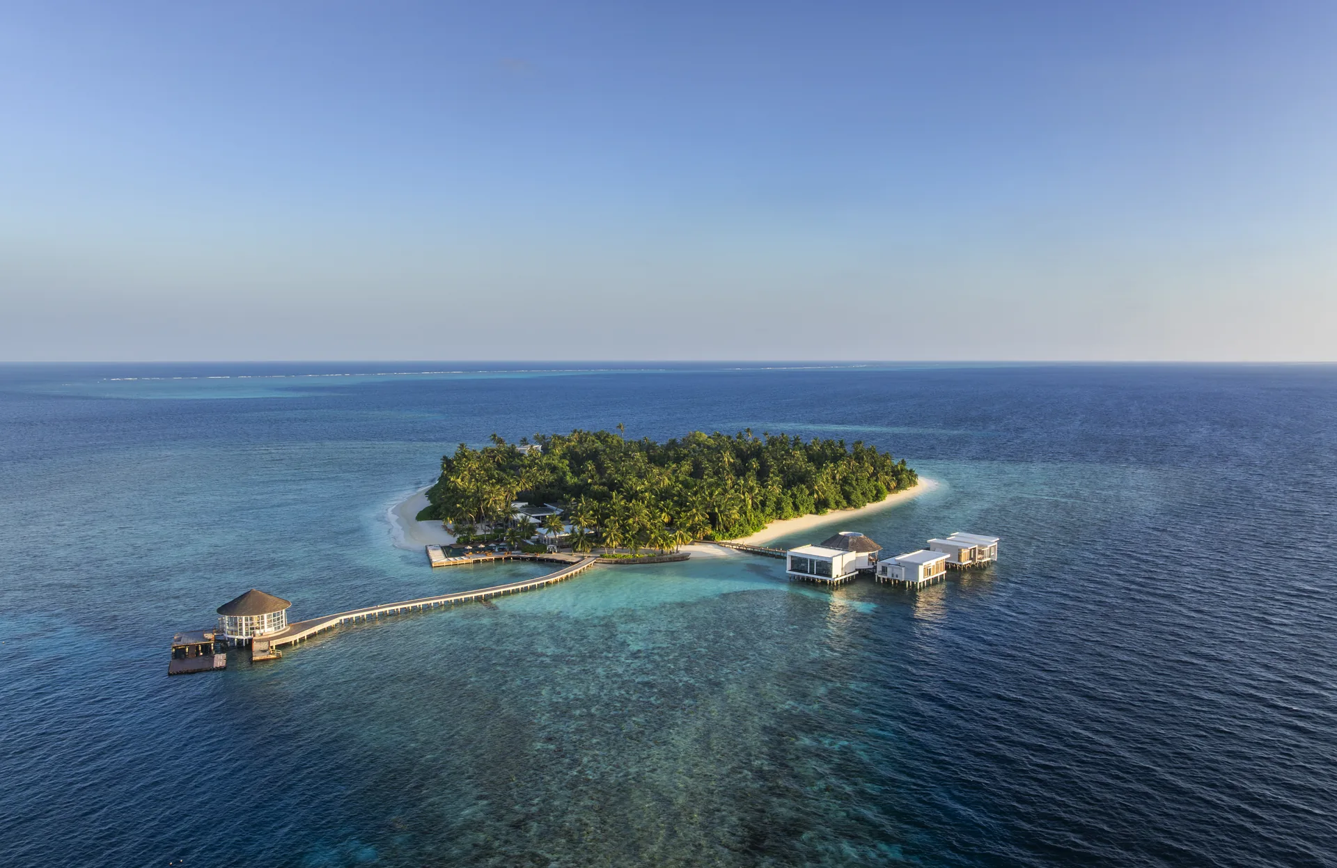 Aerial view of The Halcyon Private Isles main island in Gaafu Alifu Atoll, a green island with a white beach, long jetty and overwater villas in a deep blue sea