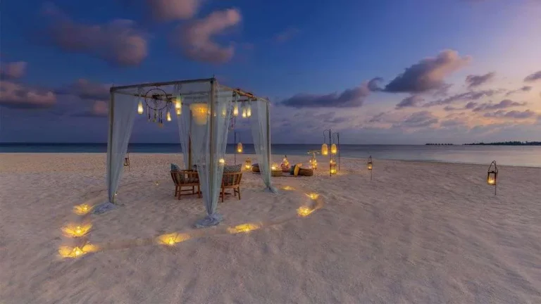 Candlelit dinner table under a white canopy on the beach at dusk, with lanterns in the sand, at Kudadoo Maldives Private Island, Lhaviyani Atoll