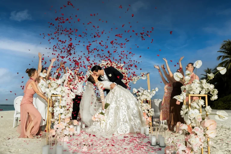 Bride and groom kiss under a shower of rose petals at a white floral beach wedding set-up with guests celebrating, Velaa Private Island, Noonu Atoll
