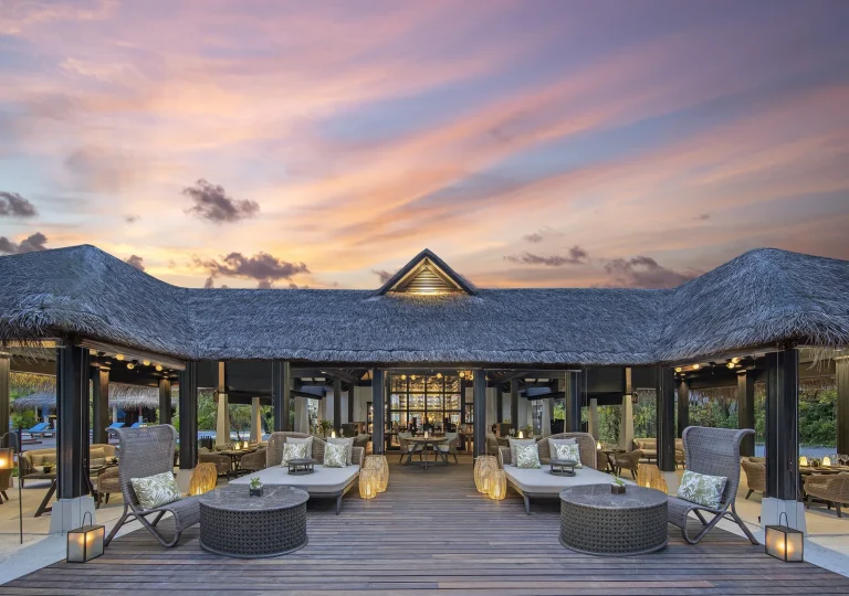 The Living Room at Naladhu Private Island at sunset, a thatched open-sided pavilion with sofas and a wooden deck facing the lagoon, South MalΓ© Atoll