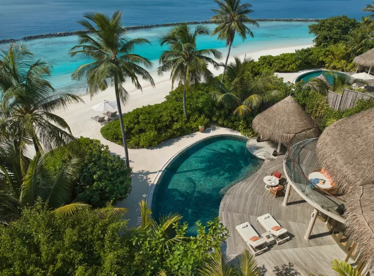 Aerial view of a thatched beach residence with a large curved pool among palms at The Nautilus Maldives, Baa Atoll