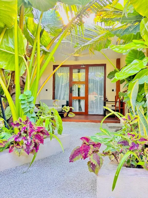 Tropical-garden path to a glass-door room with wooden steps, potted purple-pink plants and palm trees at Raakani Dhigurah, Dhigurah, Maldives