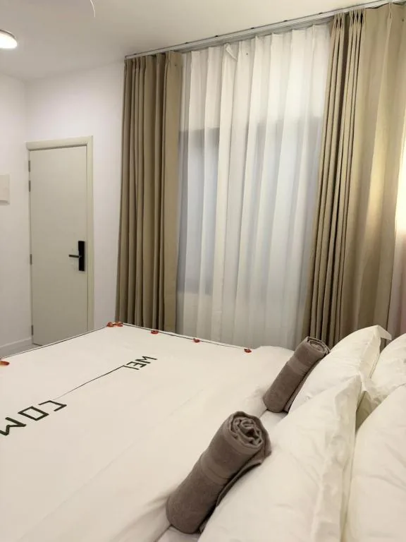 Bed with white linens and 'WELCOME' runner, two rolled towels on bed, floor-to-ceiling curtains and open door at Ranveli Dhigurah, Dhigurah, Maldives