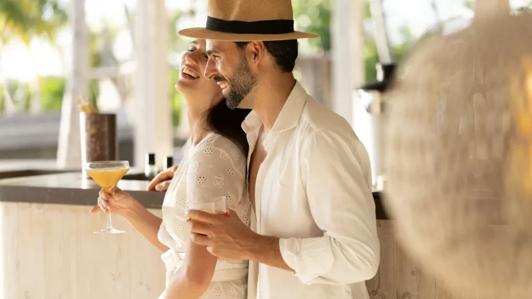 Couple laughing with cocktails at the bar of a beachfront shack at The Ritz-Carlton Maldives, Fari Islands