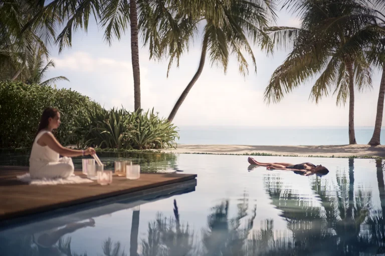 Guest floating in an infinity pool beside a therapist with singing bowls under palm trees at The Ritz-Carlton Maldives, Fari Islands