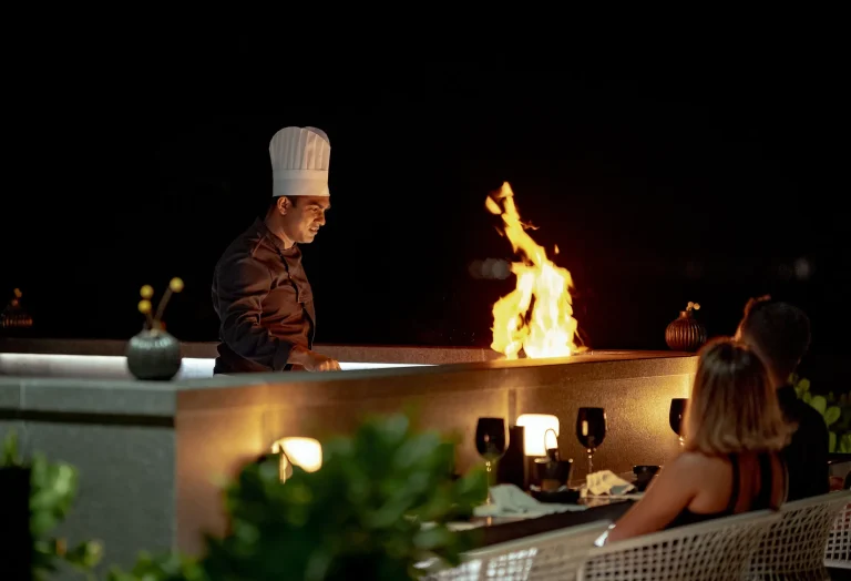 Chef cooking over flames at an open counter under the night sky for diners at IWAU, The Ritz-Carlton Maldives, Fari Islands