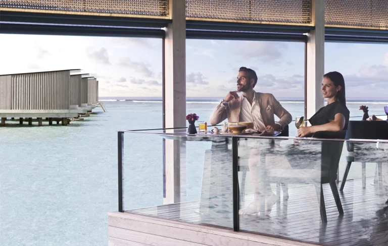 Couple dining at a window table over the lagoon with overwater villas beyond at Summer Pavilion, The Ritz-Carlton Maldives, Fari Islands
