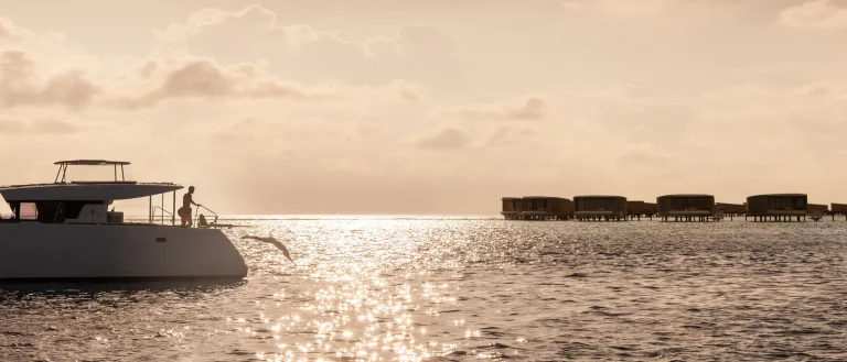 Catamaran on a glittering golden sea at sunset with overwater villas on the horizon, The Ritz-Carlton Maldives, Fari Islands