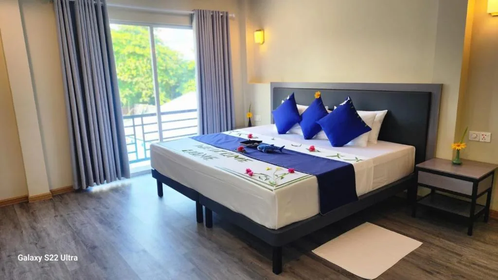 King bed with 'WELCOME HOME' runner and blue pillows, balcony access, bedside table and yellow flowers; Galaxy S22 Ultra at Seaside Dhangethi Inn, Dhangethi, Maldives