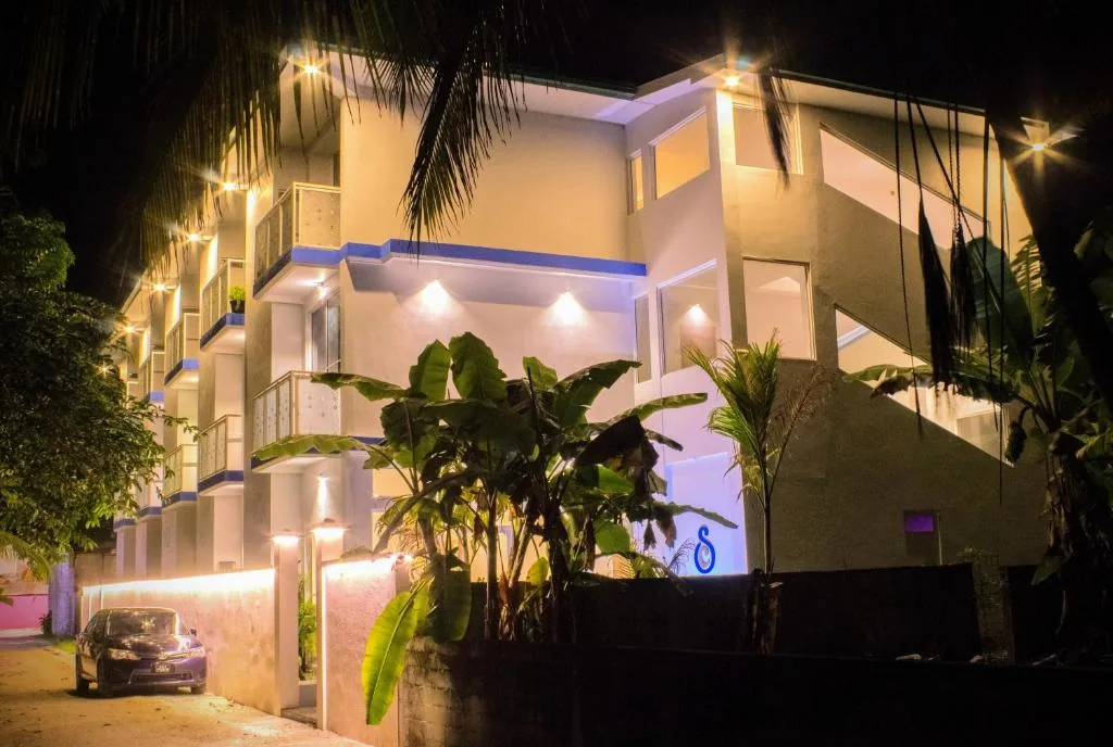 Three-story guesthouse with blue trim, recessed wall lights, palm trees and on-site parking at night at Silver County Hotel, Fuvahmulah, Maldives
