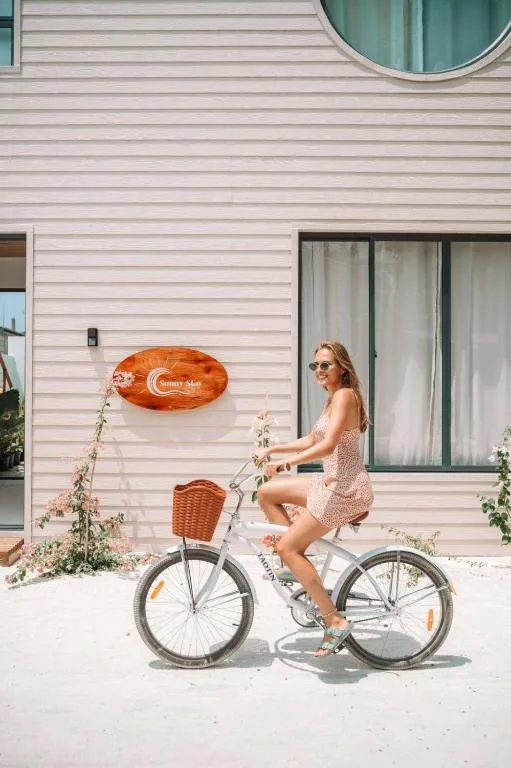 Guest riding a white bicycle with wicker basket outside a white guesthouse with 'Sunny Stay' sign and flowering trellis at Sunny Stay Dhigurah, Dhigurah, Maldives