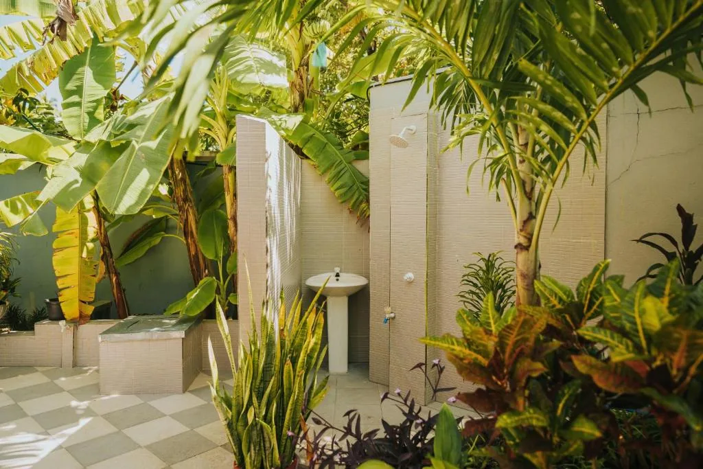 Outdoor shower with wall-mounted showerhead and sink, surrounded by banana trees and tropical plants on a checkered patio at The Sandscape, Fuvahmulah, Maldives
