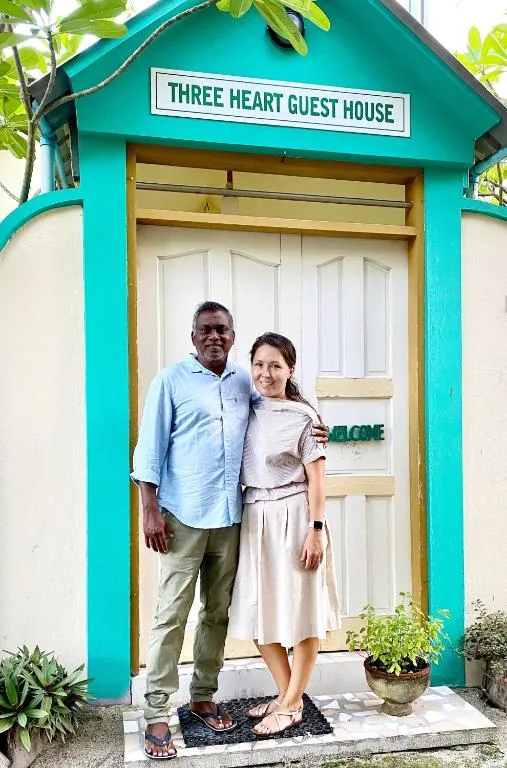 Two guests outside turquoise guesthouse with teal door labeled THREE HEART GUEST HOUSE, potted plants and 'WELCOME' sign at Three Hearts Guesthouse and Villas, Fulhadhoo, Maldives