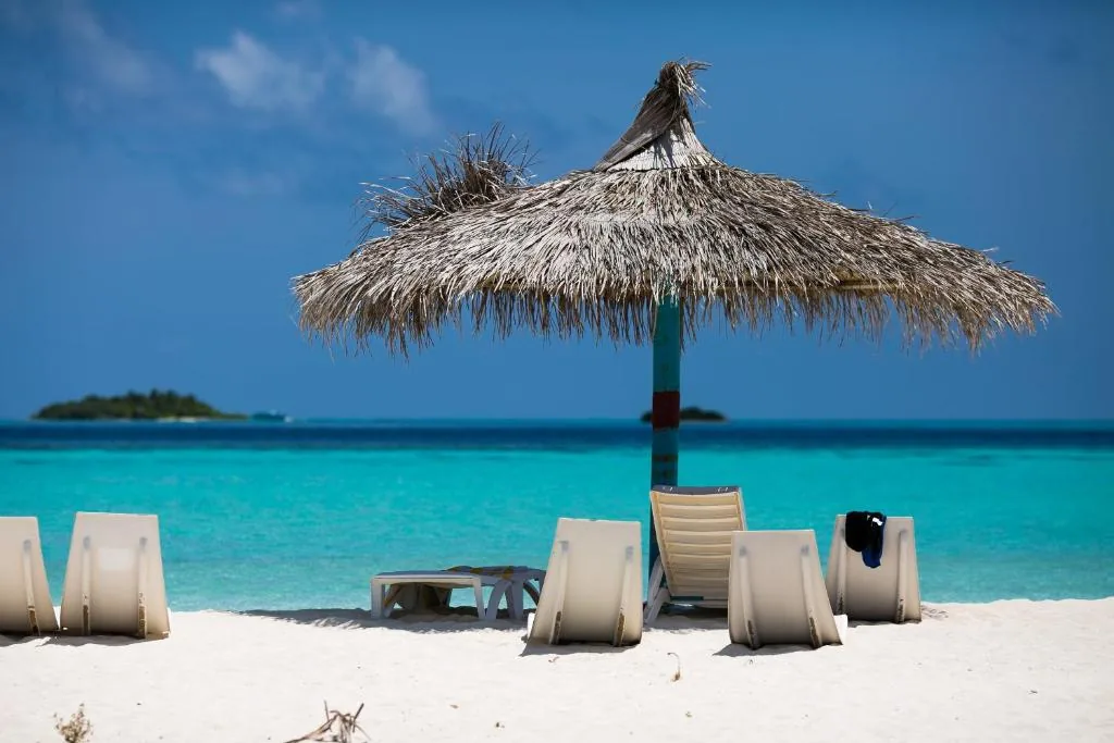 Sandy beach with a thatched palapa and white loungers by the lagoon at Triton Beach Hotel & Spa, Maafushi, Maldives