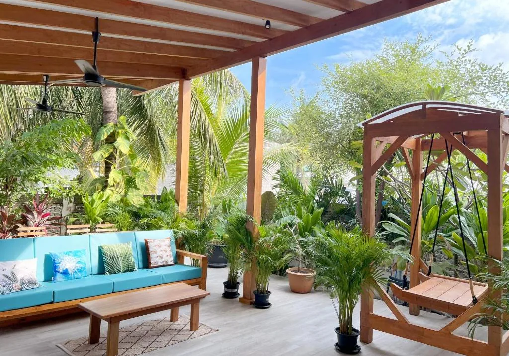 Covered wooden deck with blue sofa, ceiling fan, potted palms and hanging wooden swing in tropical garden at Villa Laguna Maldives, Fulhadhoo, Maldives